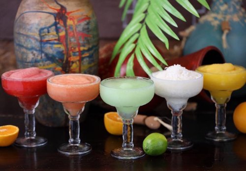 Five colorful frozen cocktails in glasses, with a tropical backdrop featuring fruits and decorative elements.