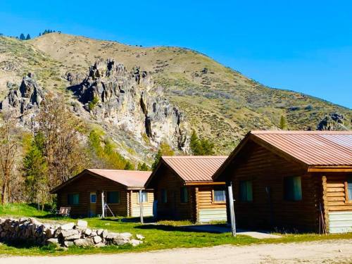 Three log cabins with metal roofs nestled against a rocky hillside under a clear blue sky.