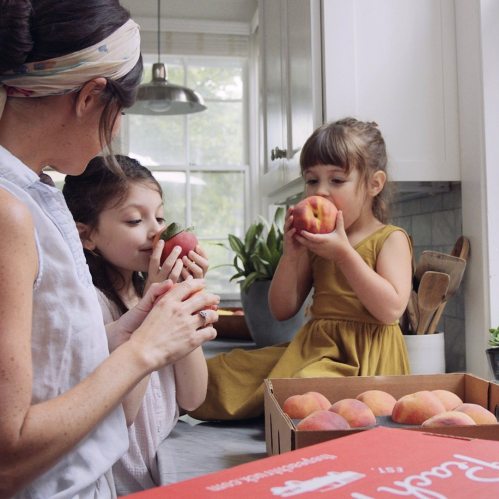 A woman and two girls enjoy fresh peaches in a bright kitchen, with one girl sitting on the counter.