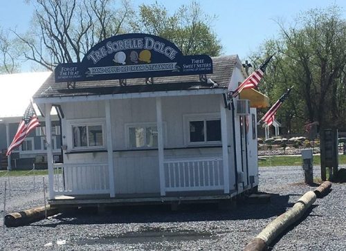 A small white kiosk with American flags, surrounded by gravel and logs, on a sunny day.