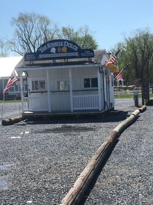 A small white kiosk with American flags, surrounded by gravel and logs, on a sunny day.