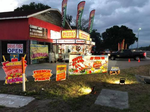 A colorful food stand with signs for pizza, pretzels, and lemonade, set against a cloudy evening sky.