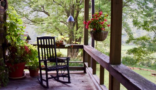 A cozy porch with a rocking chair, potted plants, and hanging flowers, surrounded by lush greenery.