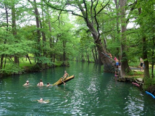 A serene river scene with people swimming, a wooden boat, and lush green trees surrounding the water.