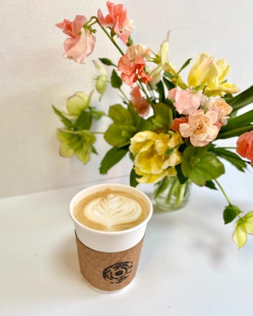 A latte in a paper cup sits beside a vase of colorful flowers on a white surface.