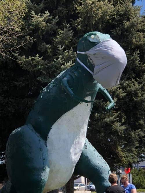 A large dinosaur statue wearing a face mask, surrounded by trees and people in the background.