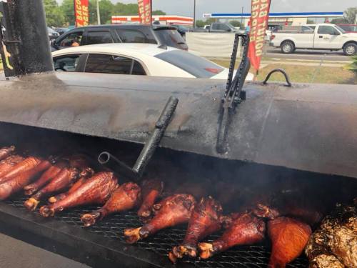 Smoked turkey legs cooking on a barbecue grill, with smoke rising and cars in the background.