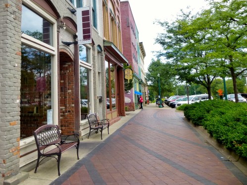 A charming street lined with shops, benches, and trees, featuring a brick walkway and parked cars in the background.