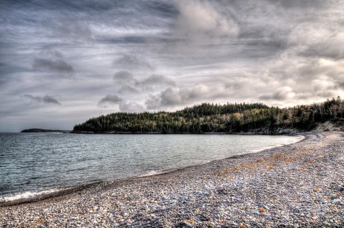 A rocky beach with calm waters, surrounded by lush green hills under a cloudy sky.
