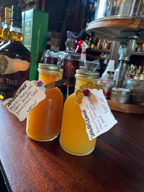 Two glass bottles with colorful drinks, labeled with tags, sitting on a bar counter surrounded by various spirits.