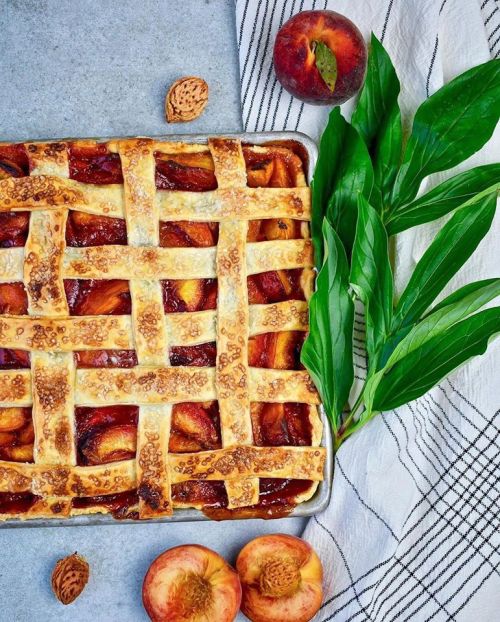 A freshly baked peach pie with a lattice crust, surrounded by fresh peaches and green leaves on a textured surface.
