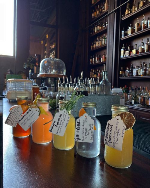 Five colorful bottles of cocktails lined up on a bar, each with a handwritten label, against a backdrop of shelves filled with spirits.