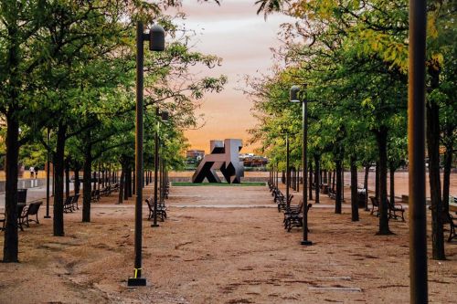 A park pathway lined with trees and benches, featuring a large abstract sculpture at the center against a sunset sky.