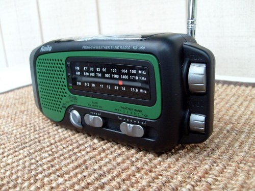 A portable weather band radio with a green and black design, featuring a dial and antenna, placed on a textured surface.