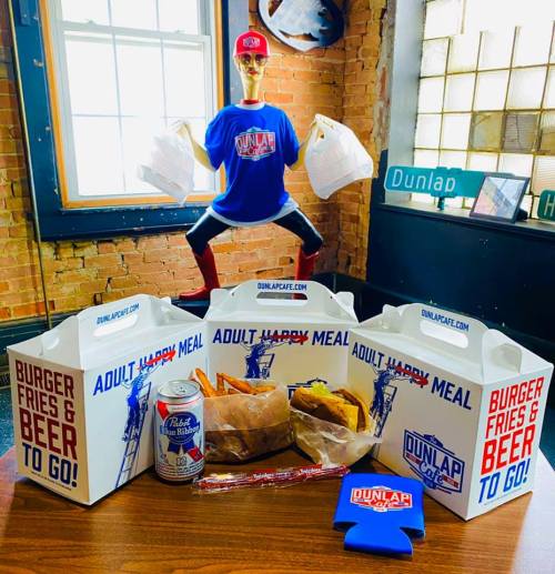A table with takeout boxes labeled "Adult Happy Meal," fries, burgers, a beer, and a mannequin wearing a Dunlap shirt.