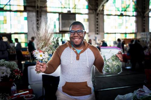 A smiling man holds a bouquet of flowers in one hand and a bag of herbs in the other, standing in a vibrant market setting.