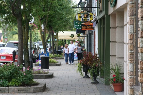 A couple walks hand-in-hand along a tree-lined sidewalk, with shops and flowers lining the path.