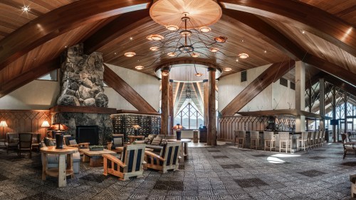A spacious lodge interior with wooden beams, a stone fireplace, cozy seating, and a bar area, featuring warm lighting.