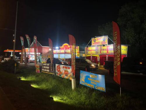 Brightly lit food stands at night, featuring funnel cakes and fried foods, with colorful banners and parked cars nearby.