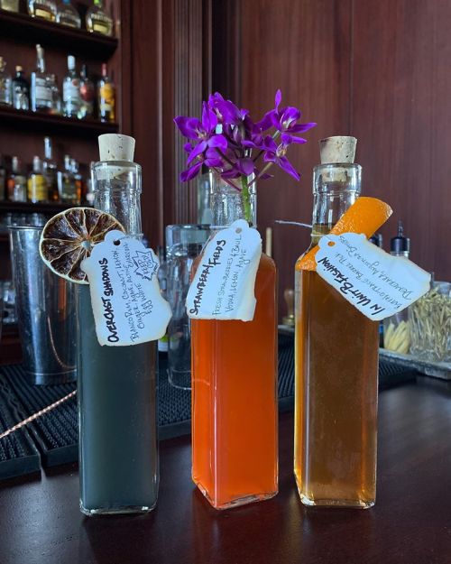 Three glass bottles with cork stoppers, each containing colorful liquids and labeled with tags, displayed on a bar counter.