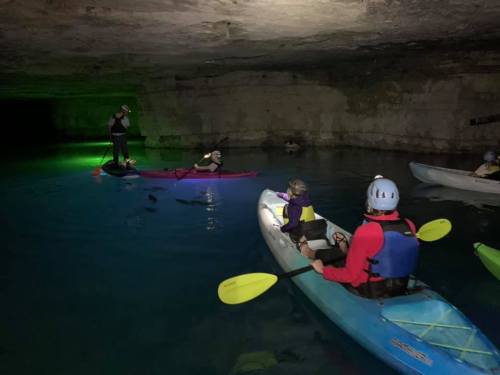 A group of kayakers paddles in a dark cave with illuminated water, wearing helmets and life jackets.