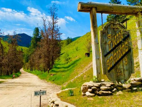 A rustic sign welcomes visitors to Twin Springs, with a dirt road and lush green hills in the background.