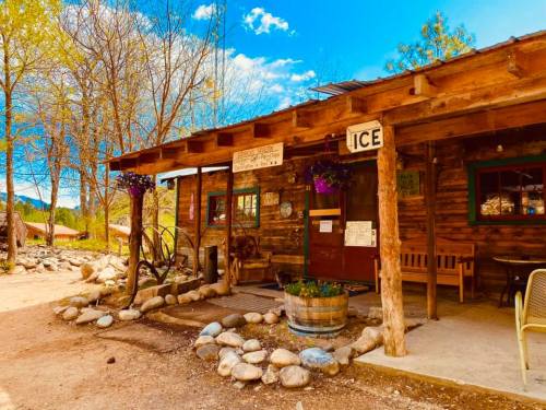 A rustic wooden cabin with a sign for "ICE," surrounded by rocks and trees under a bright blue sky.