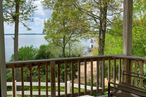 View from a porch overlooking a calm lake, surrounded by trees and greenery, with a lighthouse in the distance.
