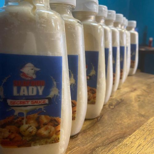 Bottles of Seafood Lady secret sauce lined up on a wooden surface, with a blue background.