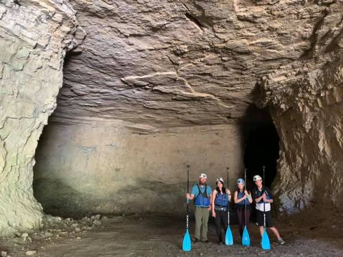 Four people stand inside a cave, holding paddles, with rocky walls and a dark opening in the background.