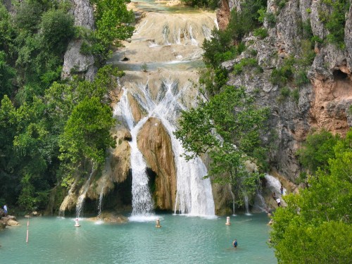 A scenic waterfall cascading over rocky cliffs into a turquoise pool, surrounded by lush greenery and people enjoying the water.