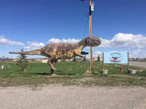 A large dinosaur statue stands beside a sign for the Two Medicine Dinosaur Center, set against a blue sky.