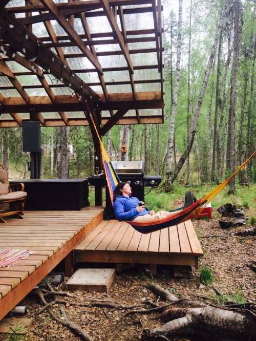 A person relaxes in a colorful hammock on a wooden deck surrounded by trees and a grill in a serene outdoor setting.