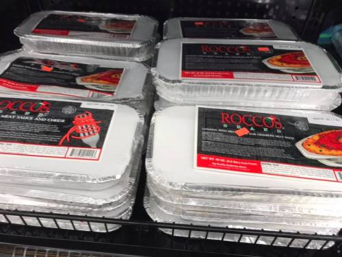 Several aluminum trays of Rocco's lasagna with meat sauce and cheese stacked on a store shelf.