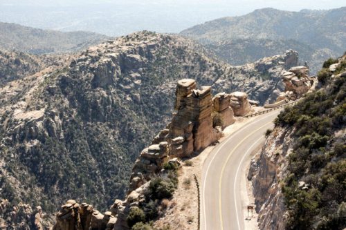 A winding road curves around rocky cliffs and mountains under a clear blue sky.