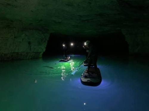 Three people paddleboarding in a dark cave with glowing blue water and headlamps illuminating their path.
