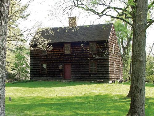 A historic wooden house with a dark exterior, surrounded by trees and a grassy area.