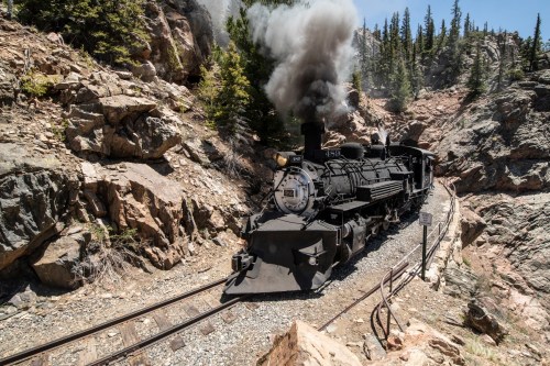 A vintage steam train chugs along rocky tracks, emitting black smoke amidst a forested mountain landscape.