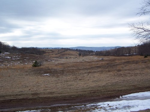 A wide, open landscape with sparse grass, patches of snow, and distant hills under a cloudy sky.