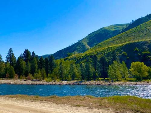 A serene river flows beside lush green hills and trees under a clear blue sky.