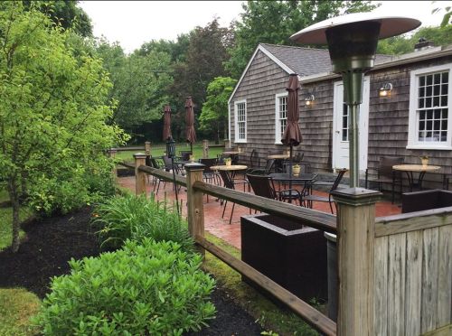 A cozy outdoor seating area with tables and umbrellas, surrounded by greenery and a rustic building in the background.