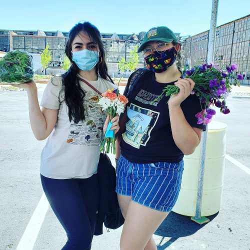 Two masked women hold bouquets of flowers and a bundle of greens, smiling in a sunny outdoor setting.