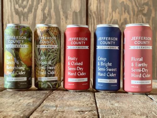 Five cans of Jefferson County Ciderworks hard cider in various flavors, displayed on a wooden surface.