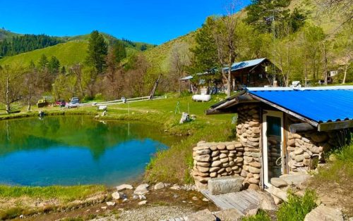 A serene landscape featuring a pond, a stone building with a blue roof, and lush green hills in the background.