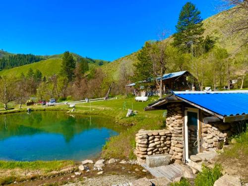 A serene landscape featuring a pond, a stone building with a blue roof, and lush green hills in the background.