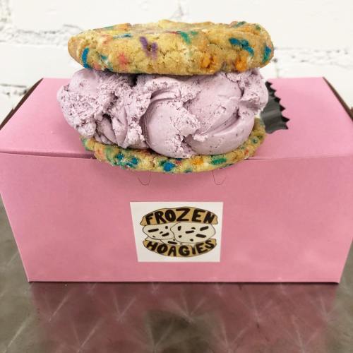 A colorful cookie sandwich filled with purple ice cream, resting on a pink box labeled "Frozen Hoagies."