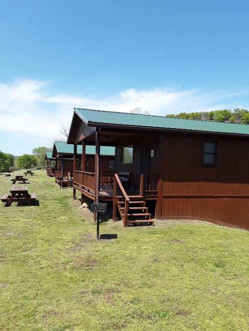 A row of brown cabins with green roofs, surrounded by grassy areas and picnic tables under a clear blue sky.