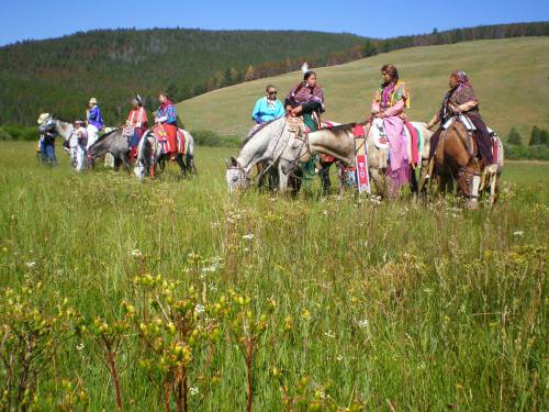 A group of people on horseback in colorful attire, standing in a lush green field with hills in the background.