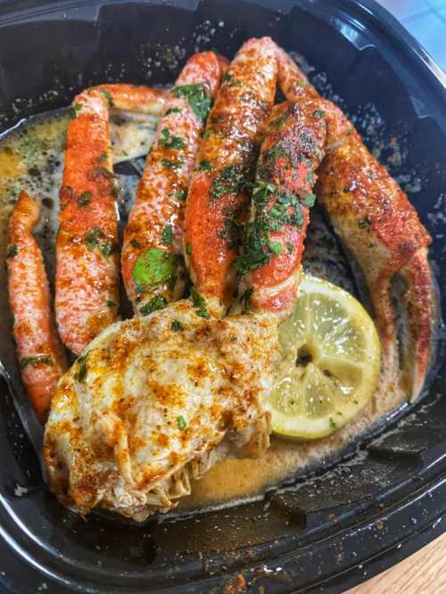 A black container filled with seasoned crab legs, a crab cake, and a lemon slice, all in a buttery sauce.
