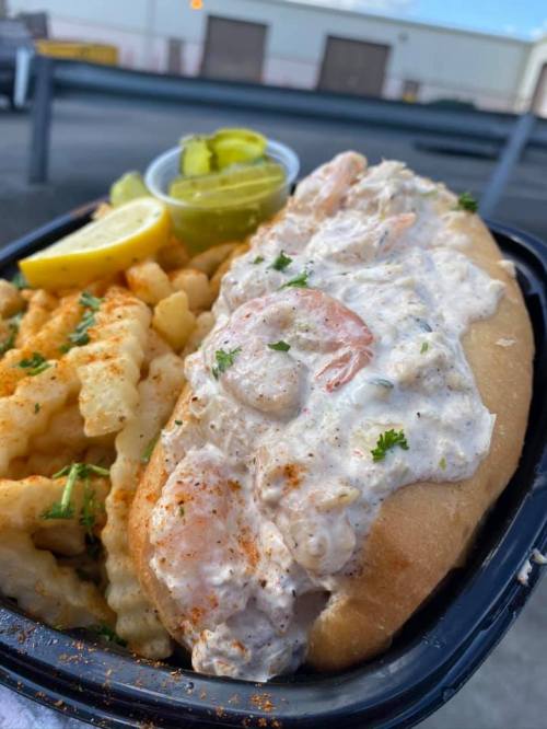 A seafood sandwich topped with shrimp and creamy sauce, served with crinkle-cut fries and pickles on the side.
